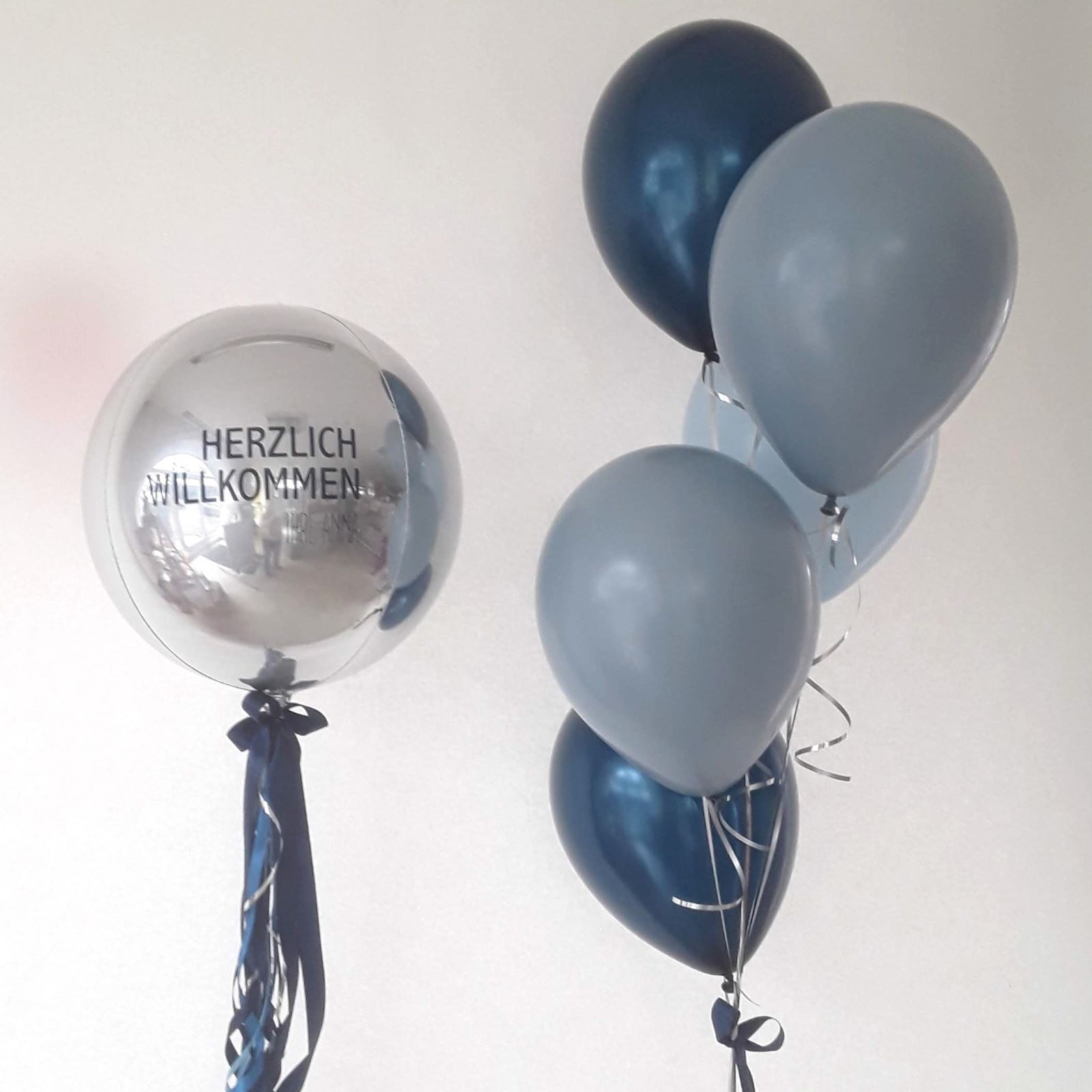 Ballonbouquet bestehend aus einem großen silbernen Orb-Ballon mit schwarzem Schriftzug „Herzlich Willkommen“ und fünf Latexballons in Dunkelblau, Hellblau und Silber, gebunden mit Bändern