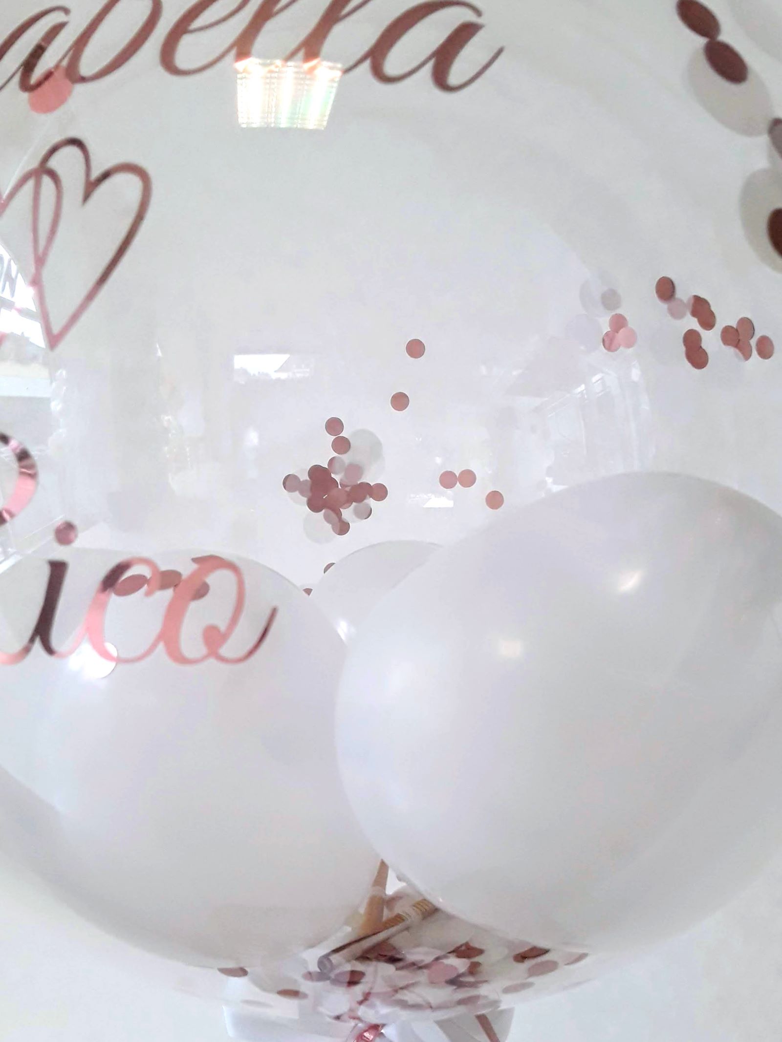 Bubbleballon mit Aufdruck 'Isabella Rico' und zwei kleinen Herzen gefüllt mit kleinen weißen Ballons, auf Ständer mit Schleifen in weiß und rosegold