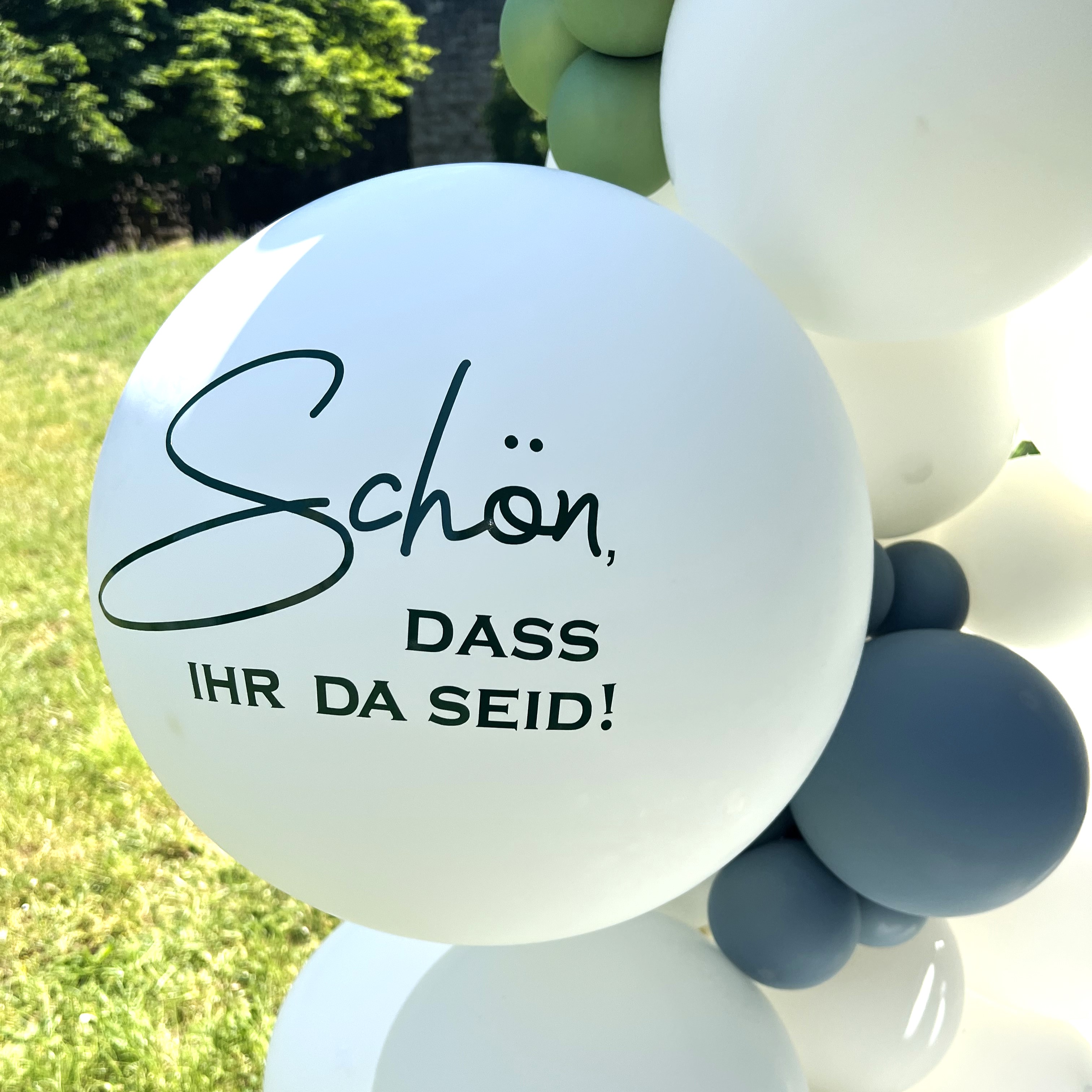 Halbkreisförmiger Ballonbogen in Weiß mit grünen und blauen Akzenten, dekoriert mit Eukalyptuszweigen und Ballon mit Schriftzug „Schön, dass ihr da seid!“3