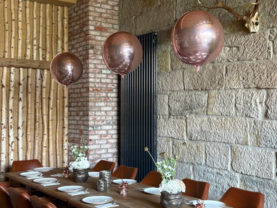 3 Schwebende Orb-Ballons in glänzendem Roségold als elegante Tischdekoration über einer festlich gedeckten Tafel in einem Raum mit Mauer- und Holzwänden