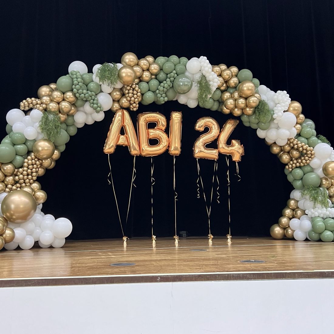 Große Ballongirlande auf Bühne in den Farben Grün, Weiß und Gold mit goldenem Schriftzug 'ABI 24' aus Folienballons, dekoriert mit Farn