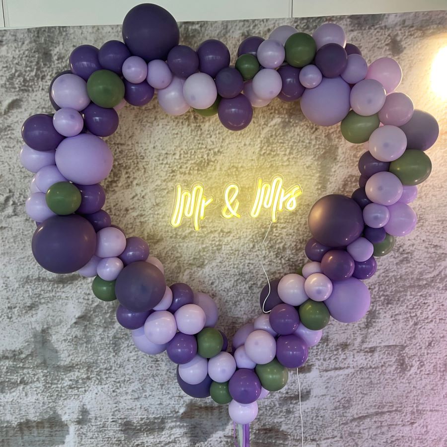 Herzförmiger Ballonbogen in vielen Farben mit unterschiedlich großen Ballons dekoriert und „Mr & Mrs“-Schriftzug in der Mitte lila
