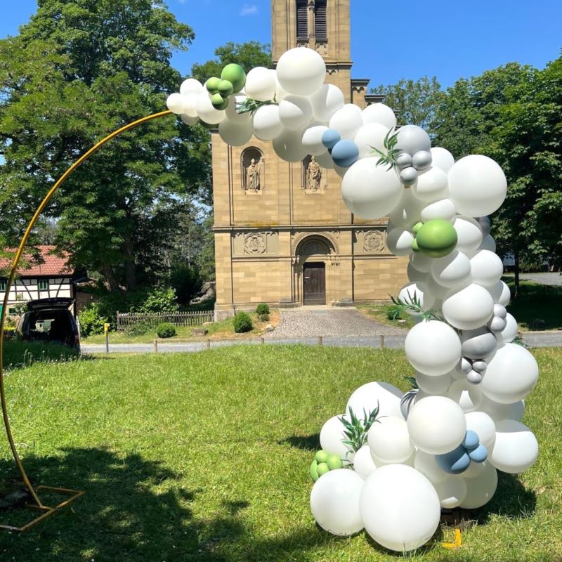Halbkreisförmiger Ballonbogen in Weiß mit grünen und blauen Akzenten, dekoriert mit Eukalyptuszweigen und Ballon mit Schriftzug „Schön, dass ihr da seid!“