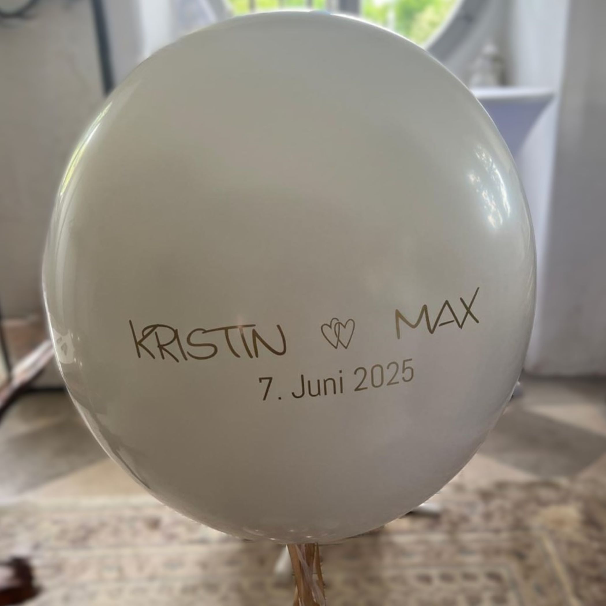 Großer weißer Heliumballon mit goldener Aufschrift 'Kristin ♥ Max 7. Juni 2025', dekoriert mit goldenen und weißen Ballonclustern am Boden.