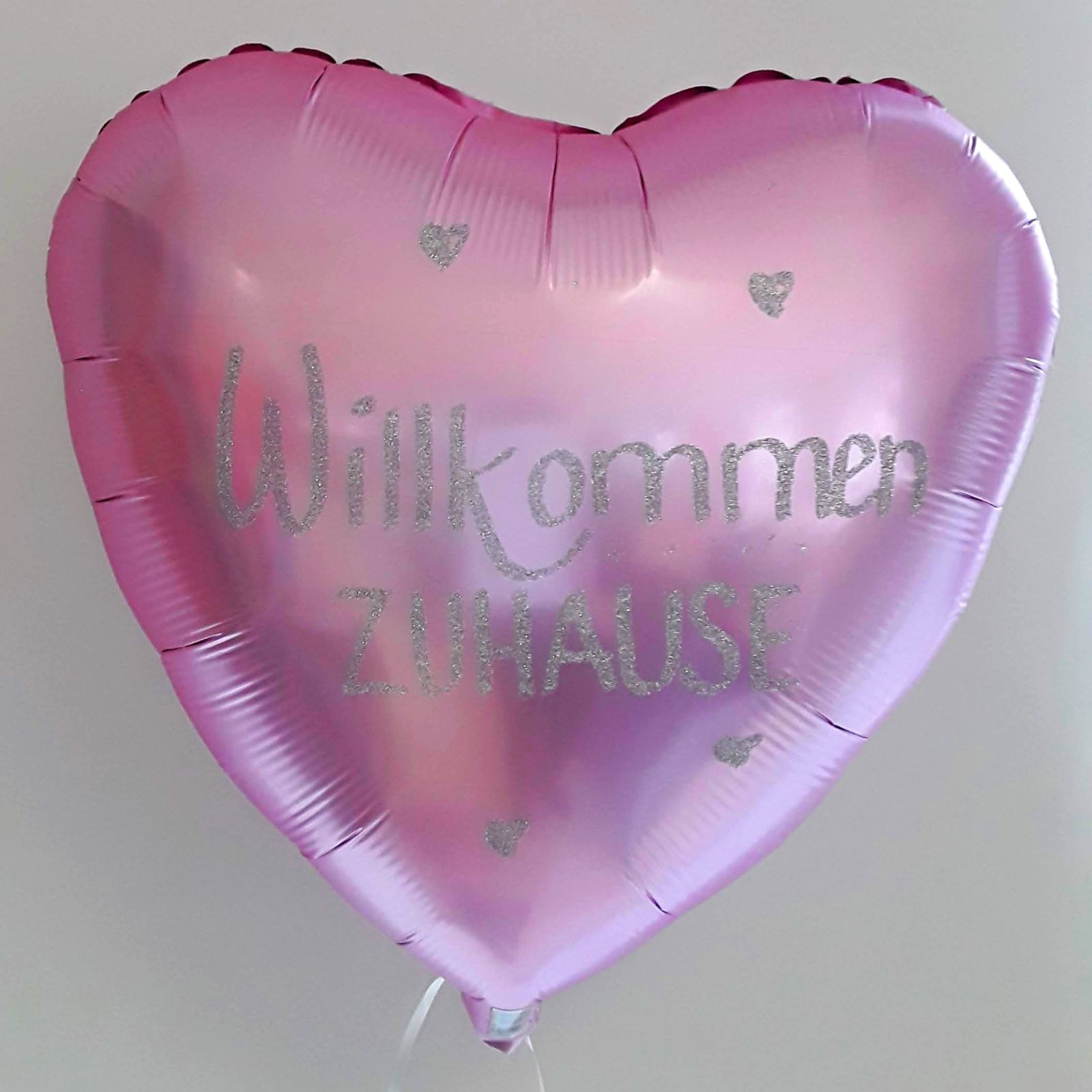 Herzförmiger Folienballon in Rosa mit silberglitzernder Aufschrift „Willkommen Zuhause“ und kleinen Herzmotiven