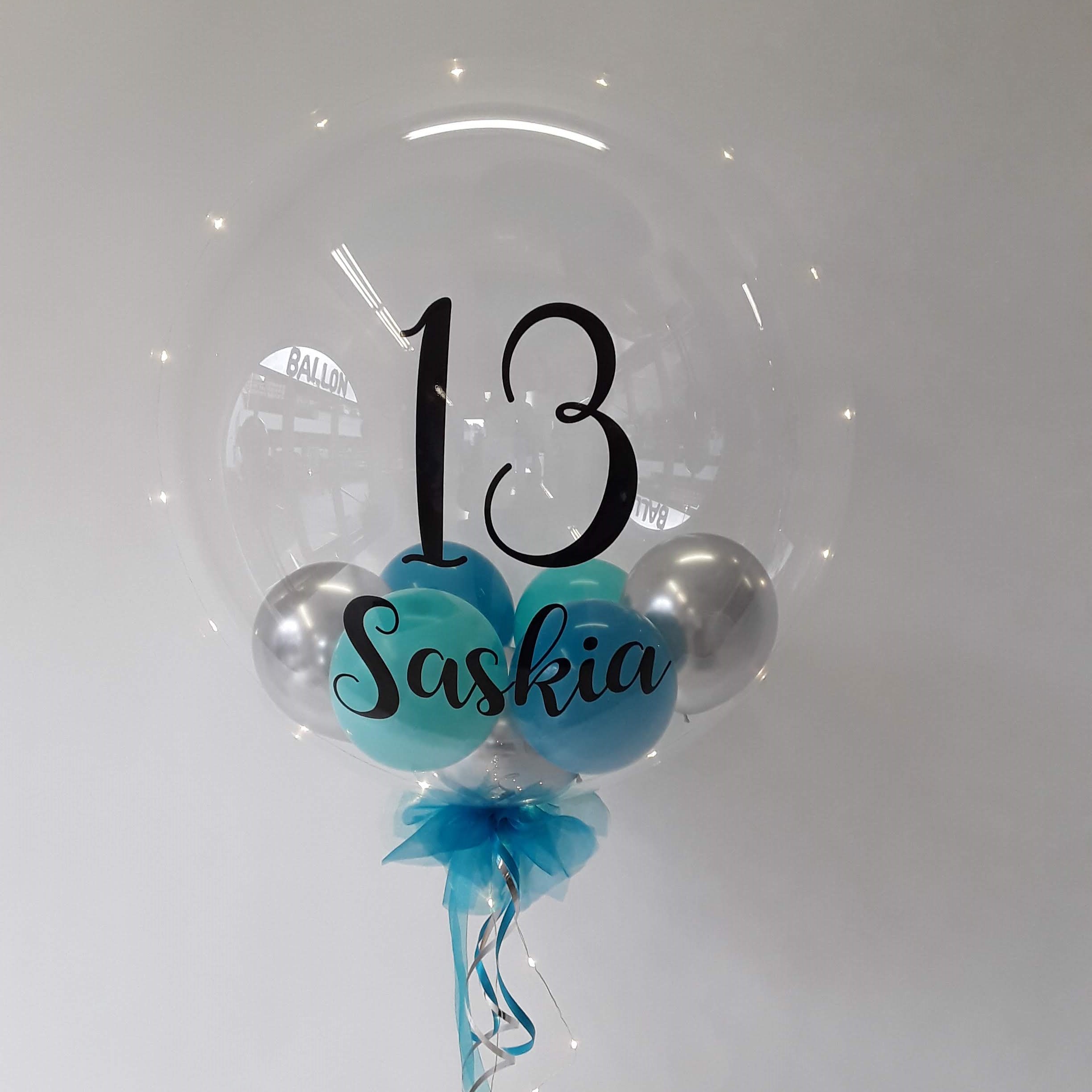 Transparenter Bubble-Ballon mit schwarzer Aufschrift „13 Saskia“, gefüllt mit silbernen und türkisfarbenen Ballons, dekoriert mit blauer Tüllschleife, silbernem Band und integrierter Lichterkette.