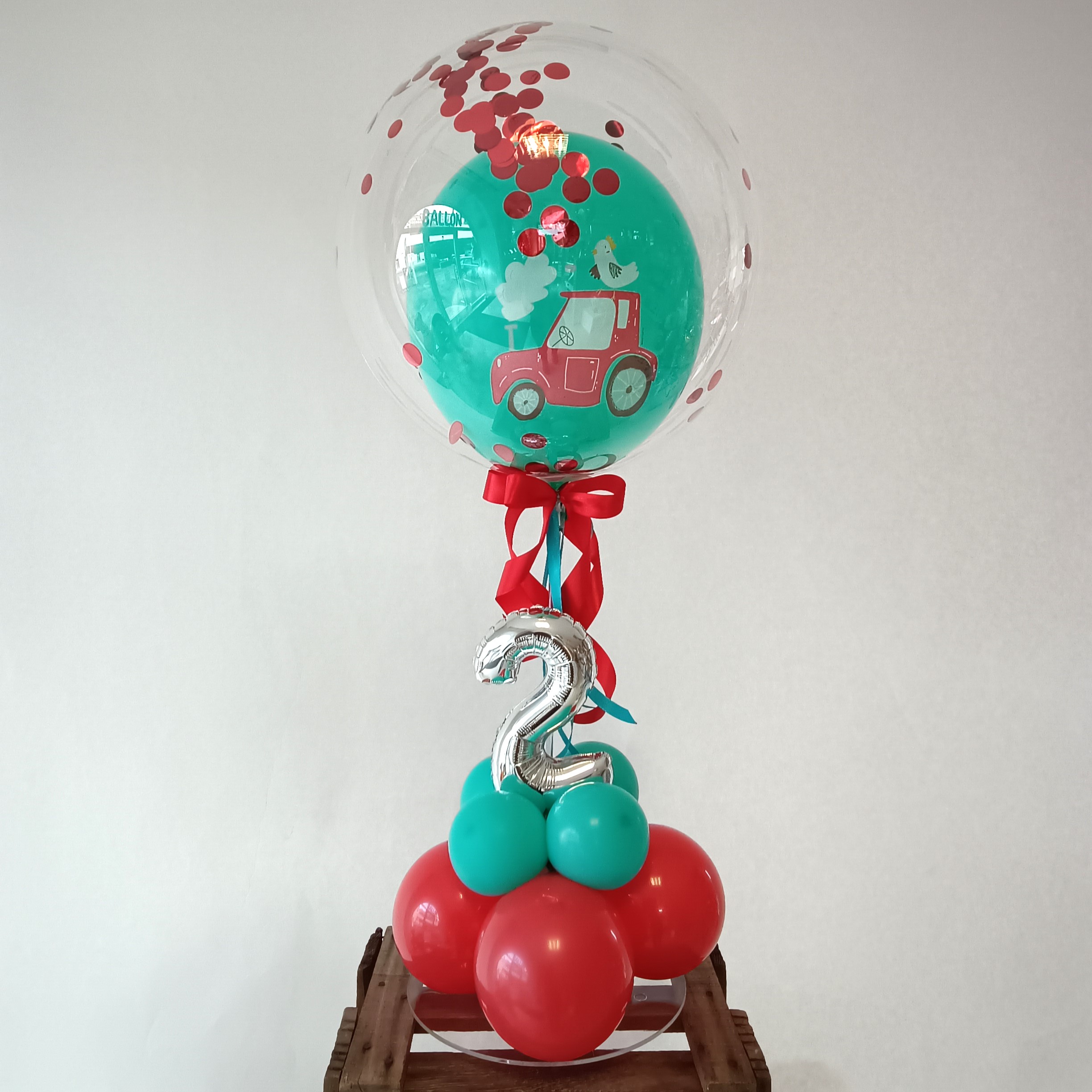 Ballondekoration mit durchsichtiger Kugel gefüllt mit rotem Konfetti, innen ein Ballon mit Autoaufdruck, kleine silberne Zahl 2, auf rotem-türkisem Ballon-Sockel.