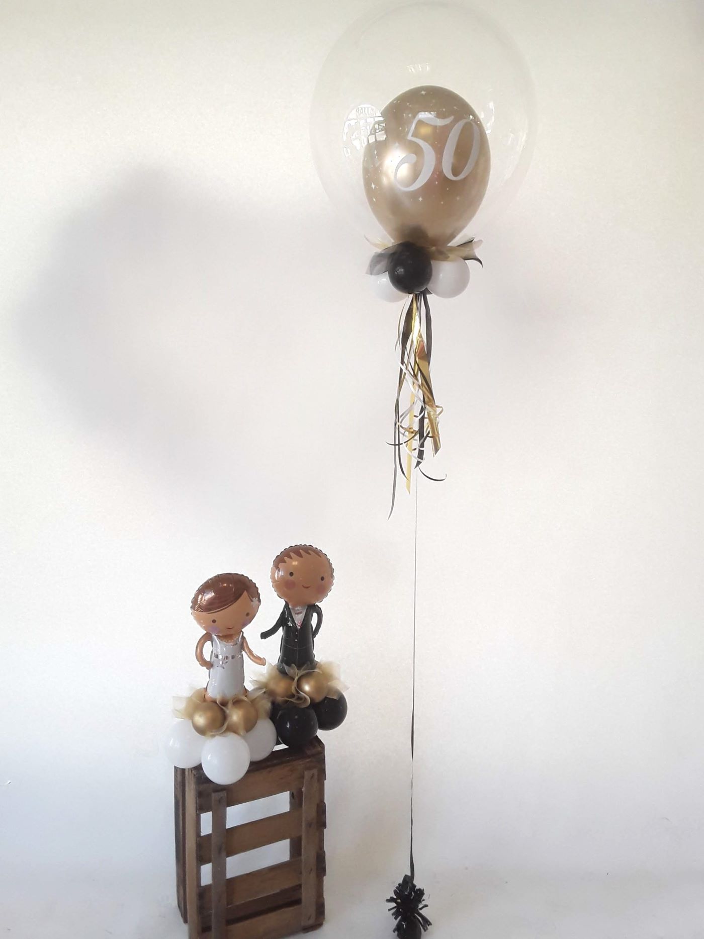 Transparenter Bubbleballon mit goldenem Innenballon mit der Zahl 50; daneben eine Brautpaar-Dekoration auf Ballonpodest in Weiß, Schwarz und Gold.