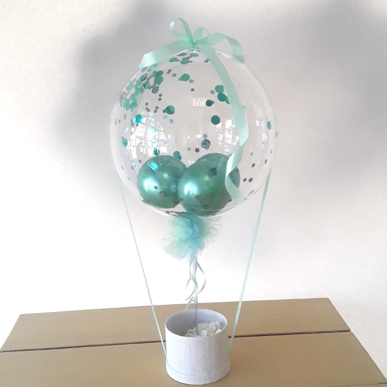 Transparenter Ballon mit türkisem Konfetti und Schleife, auf weißem Blumentopf als Basis.