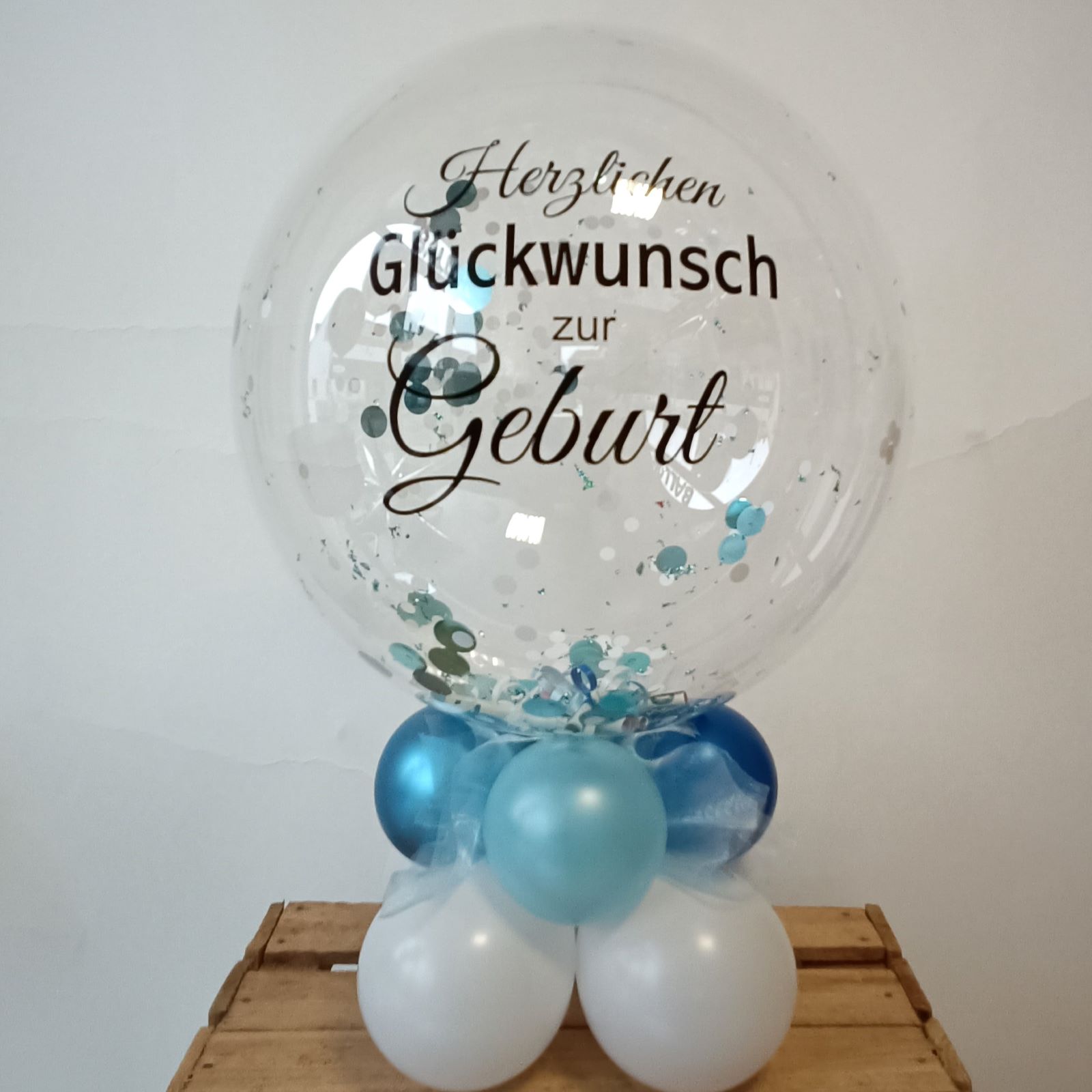 Transparenter Bubble-Ballon mit Aufdruck 'Herzlichen Glückwunsch zur Geburt', gefüllt mit blauen Konfettiballons und auf einem Sockel aus weißen und blauen Latexballons stehend