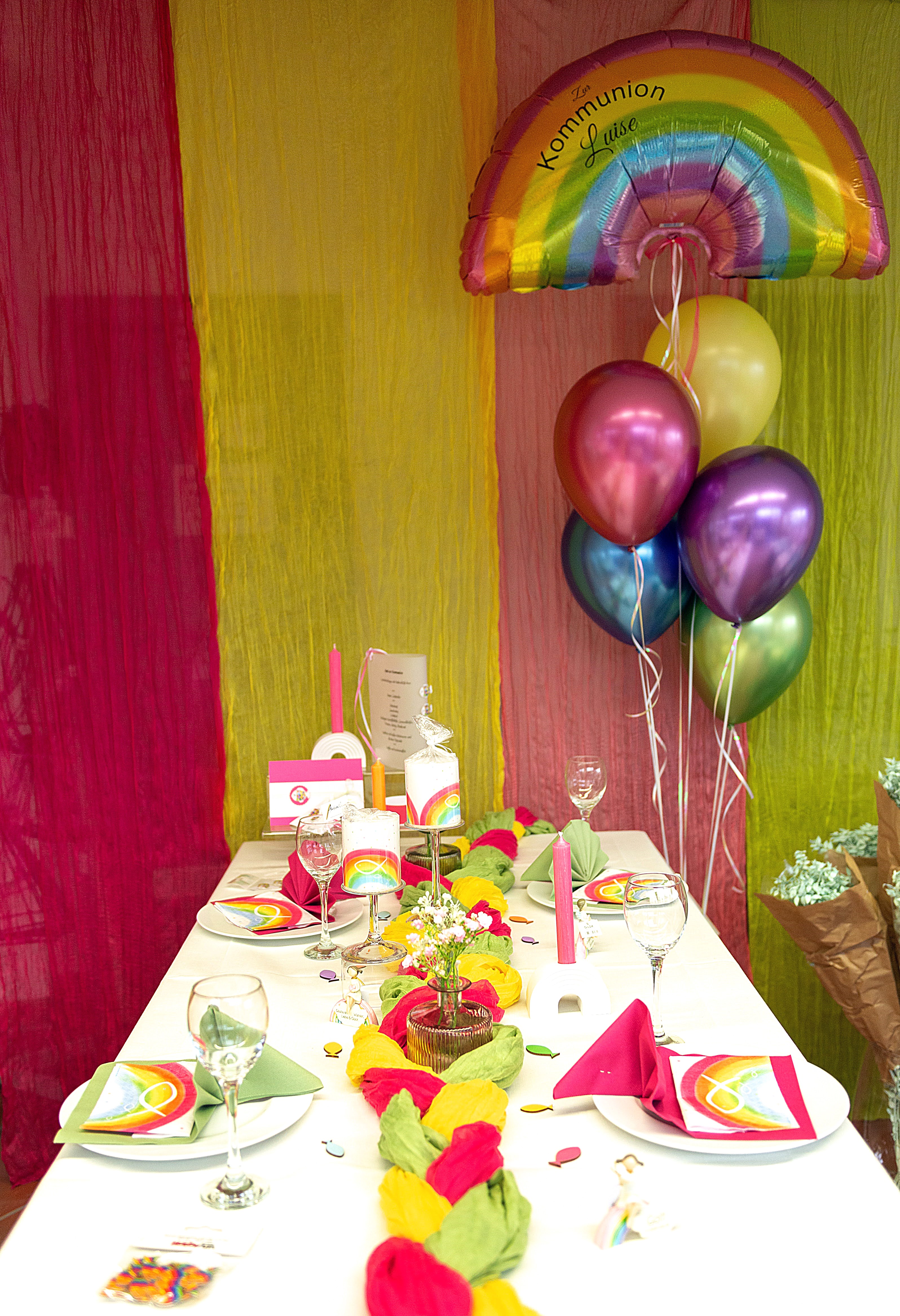 Regenbogen-Folienballon mit Aufdruck 'Zur Kommunion Luise' an einem Band mit bunten Latexballons, bunt gedeckter Tisch in den Farben rot, gelb, grün auf weißer Tischdecke