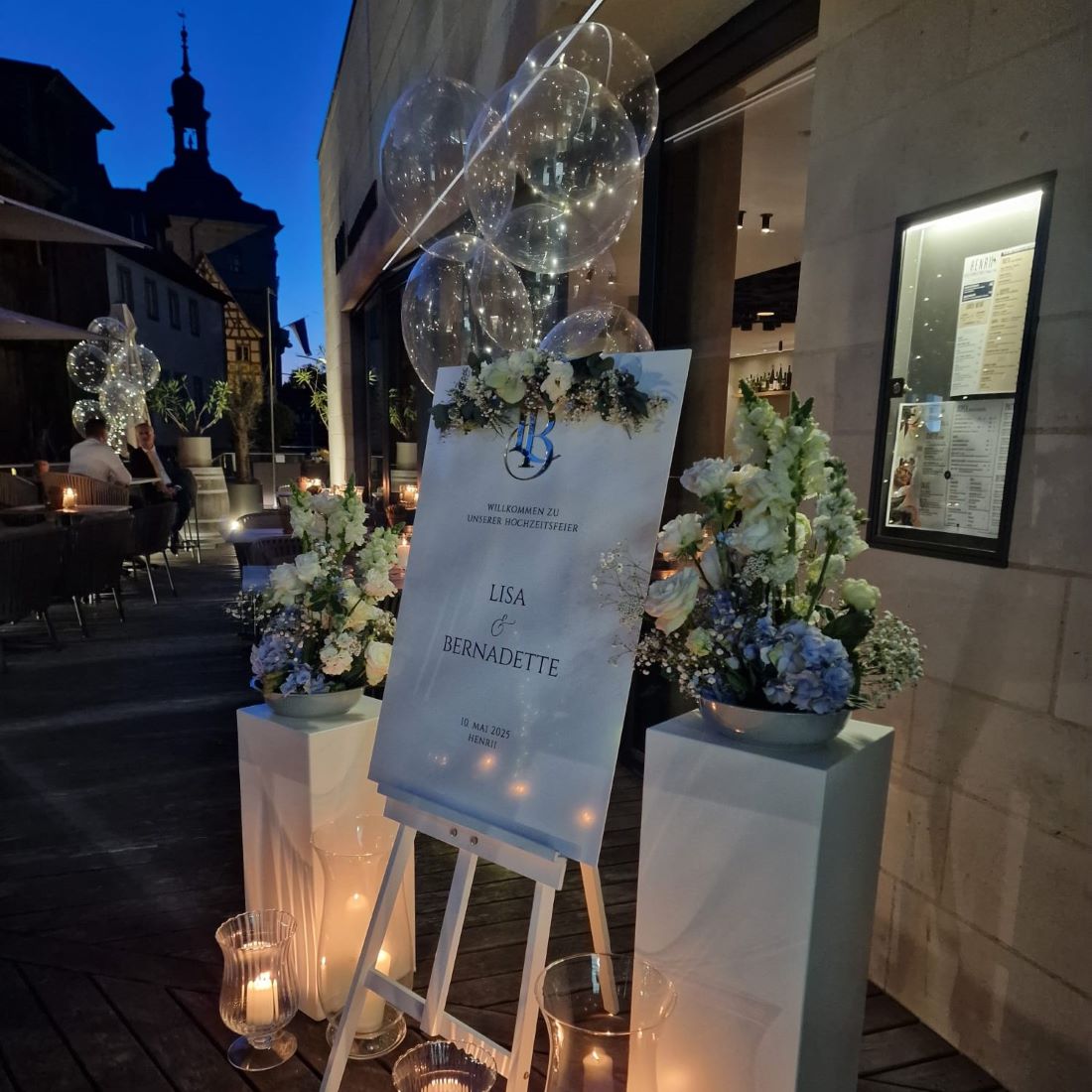 Transparente Bubble-Ballons mit integrierter Lichterkette neben einem Willkommensschild, Blumengestecken und Kerzen bei einer Hochzeit im Außenbereich bei Nacht