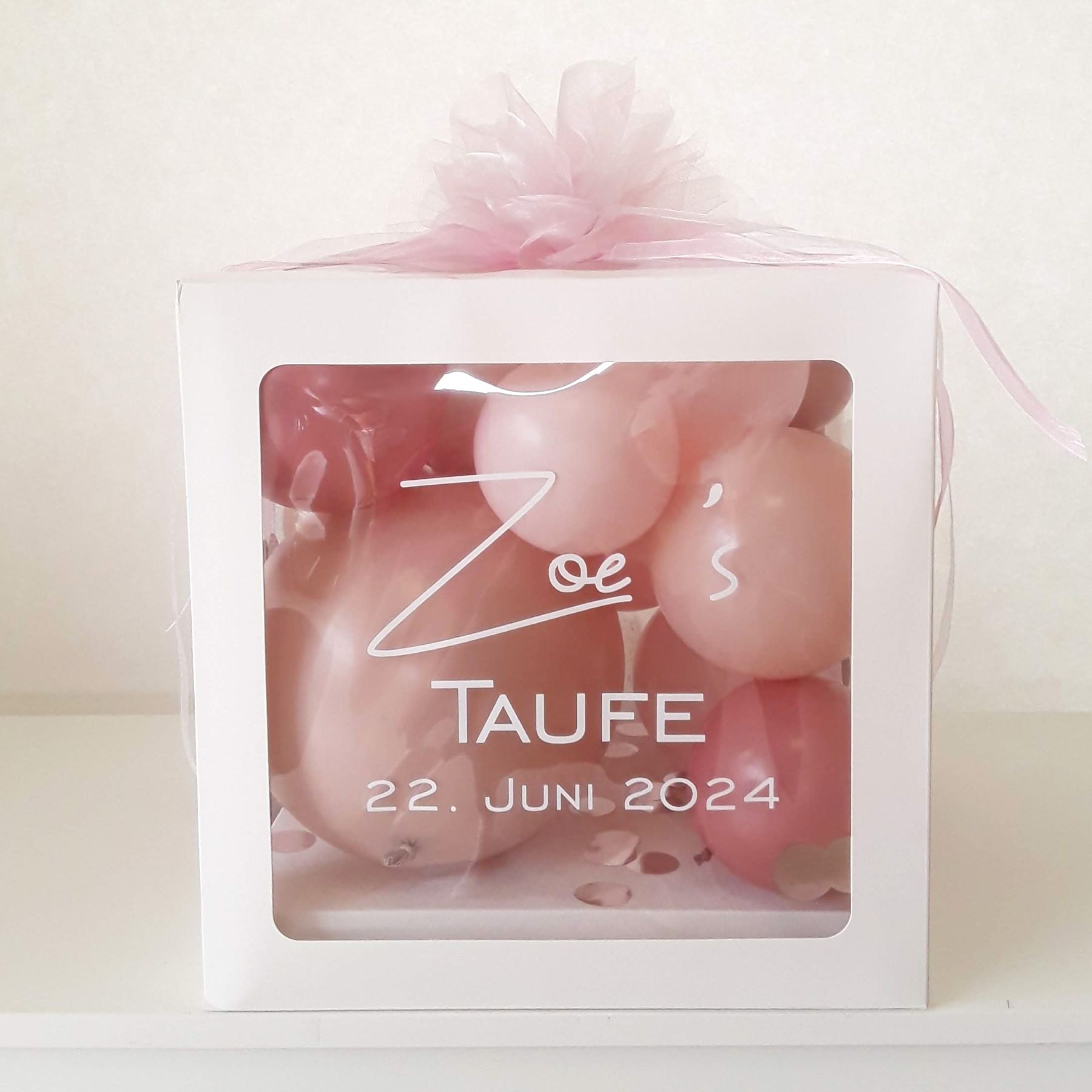 Weiße Geschenkbox mit Aufdruck 'Zoe’s Taufe – 22. Juni 2024', gefüllt mit rosa Ballons und Konfetti, dekoriert mit rosa Schleife.