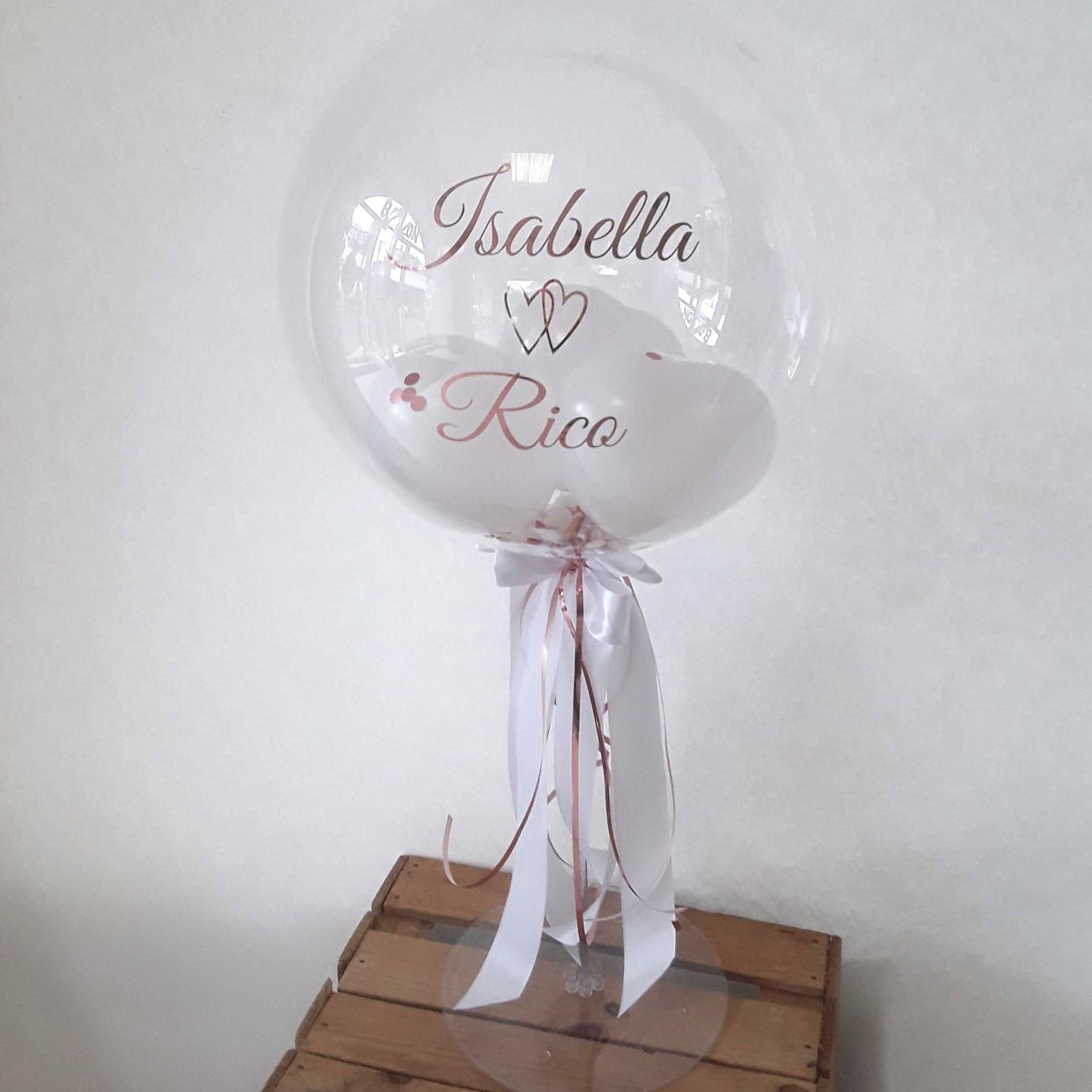 Bubbleballon mit Aufdruck 'Isabella Rico' und zwei kleinen Herzen gefüllt mit kleinen weißen Ballons, auf Ständer mit Schleifen in weiß und rosegold