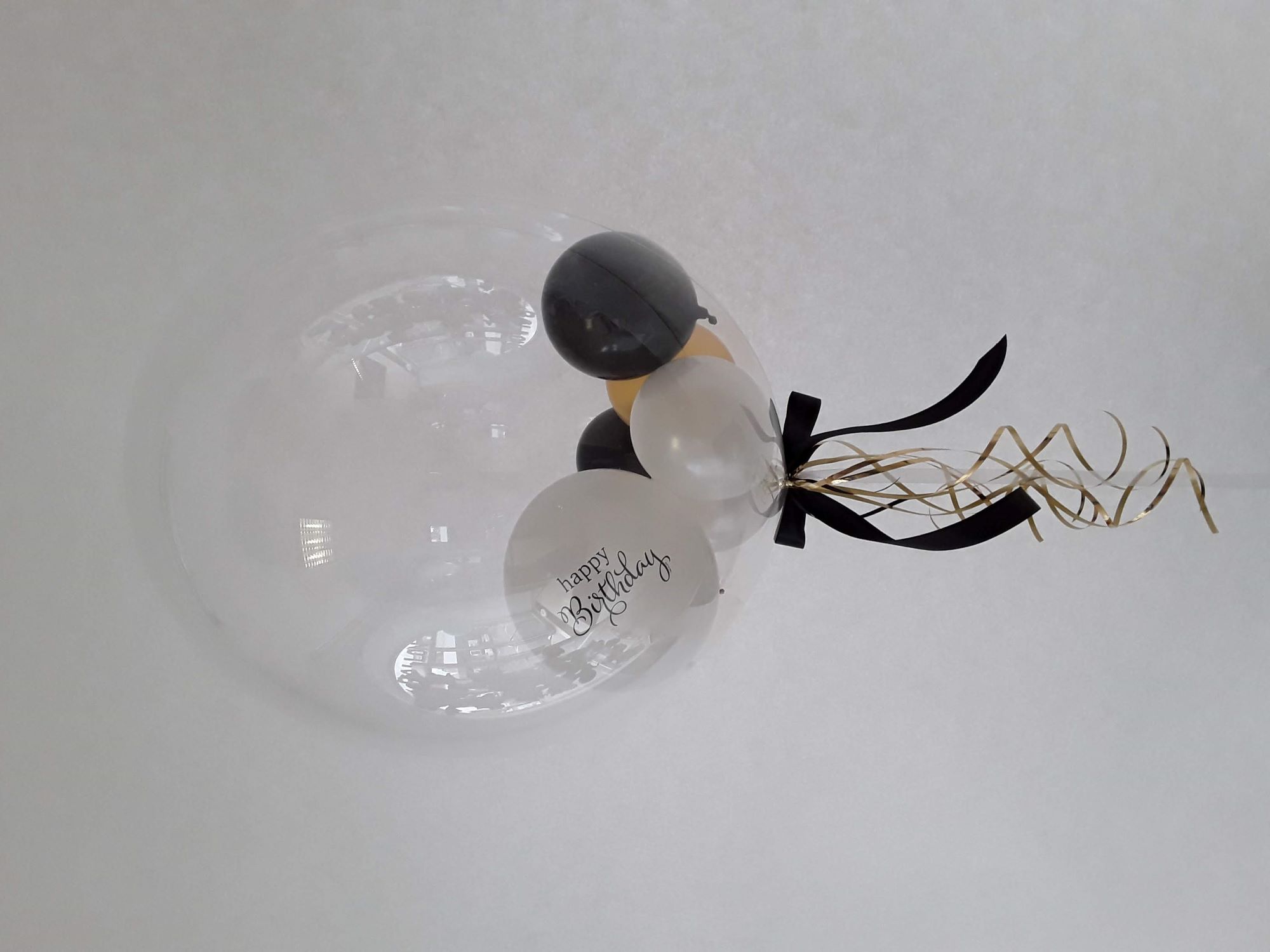 Transparenter Bubble-Ballon gefüllt mit schwarzen, weißen und goldfarbenen Mini-Ballons, davon einer mit „Happy Birthday“-Aufdruck, dekoriert mit schwarzer Schleife und goldenen Curlingbändern.