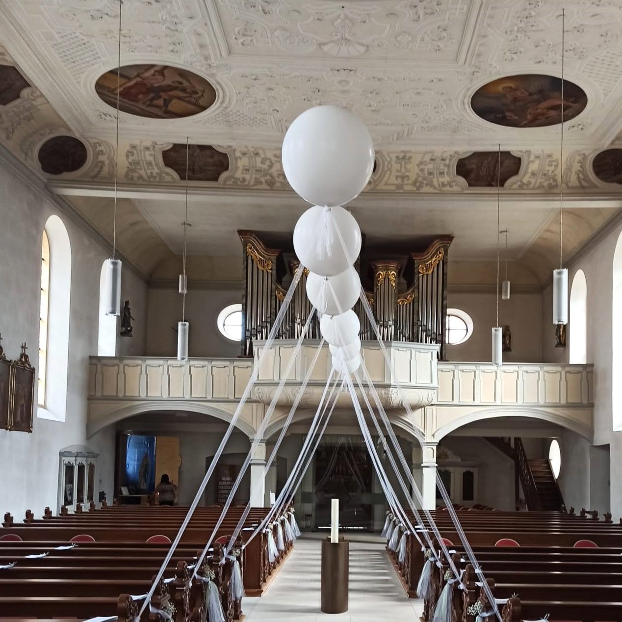 Reihe schwebender weißer Heliumballons mit Bändern, die beidseitig des roten Teppichs in einer Kirche angeordnet sind.