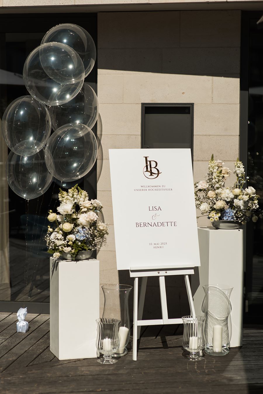 Transparente Bubble-Ballons mit integrierter Lichterkette neben einem Willkommensschild, Blumengestecken und Kerzen bei einer Hochzeit im Außenbereich.