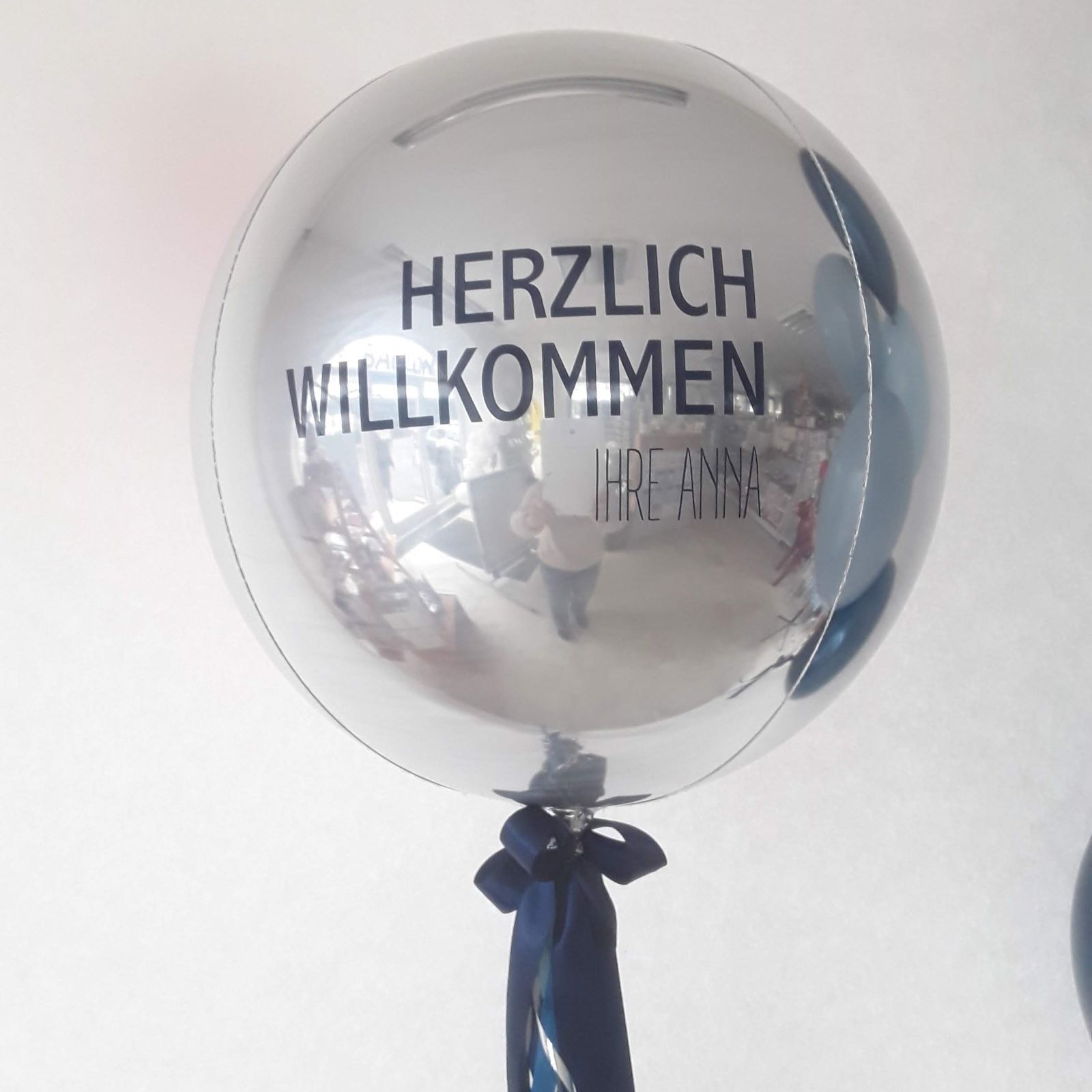 silberner Orb-Ballon mit schwarzem Schriftzug „Herzlich Willkommen Ihre Anna“ , gebunden mit Bändern