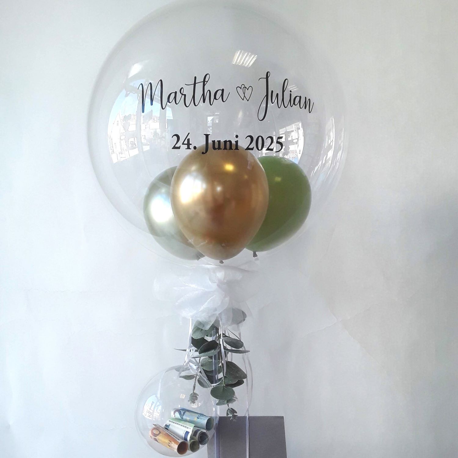 Transparenter Ballon mit Aufdruck 'Martha & Julian 24. Juni 2025' und goldenen und grünen Miniballons darin – auf Ständer mit transparentem Mini-Ballon inkl. Geldscheine, grauer Umschlag und Eukalyptus-Deko