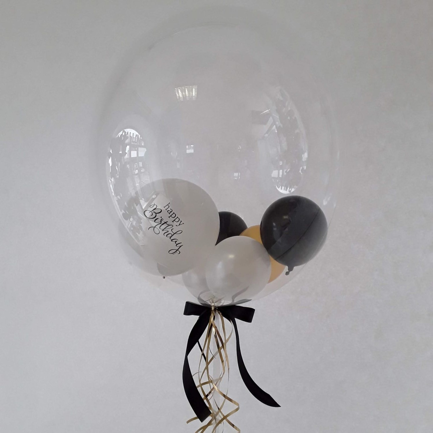 Transparenter Bubble-Ballon gefüllt mit schwarzen, weißen und goldfarbenen Mini-Ballons, davon einer mit „Happy Birthday“-Aufdruck, dekoriert mit schwarzer Schleife und goldenen Curlingbändern.