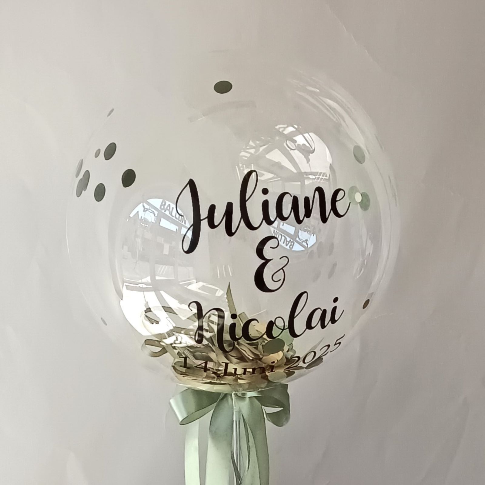 Hochzeitsballon mit Aufdruck 'Juliane & Nicolai 14. Juni 2025' gefüllt mit Geldscheinen und Konfetti auf Ständer mit Satinband in Grün