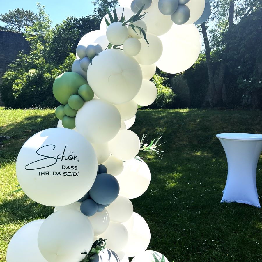 Halbkreisförmiger Ballonbogen in Weiß mit grünen und blauen Akzenten, dekoriert mit Eukalyptuszweigen und Ballon mit Schriftzug „Schön, dass ihr da seid!“4