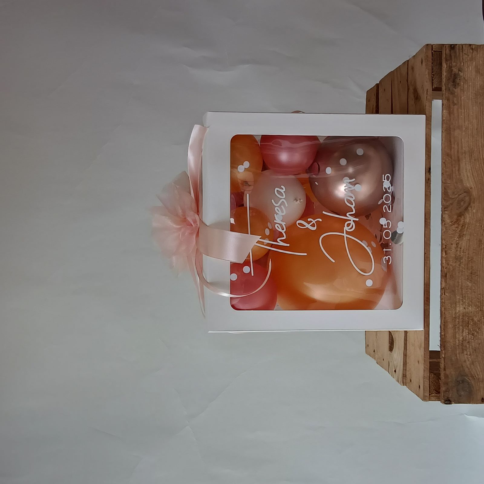 Ballonbox mit roséfarbenen, weißen und apricotfarbenen Ballons, Schleife und Tüllblume, Aufdruck mit Namen und Hochzeitsdatum.