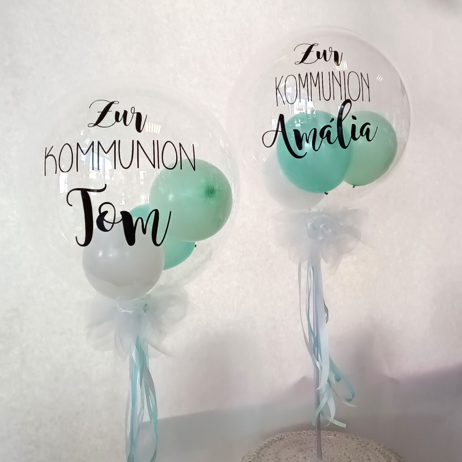 Zwei Bubbleballons mit Aufdruck 'Zur KOMMUNION Tom' und 'Zur KOMMUNION Amalia', gefüllt mit mintfarbenen und weißen Ballons, weiße Schleifen