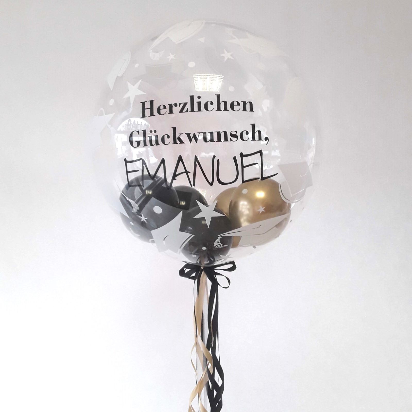 Transparenter Bubble-Ballon mit weißen Graduation-Symbolen und personalisiertem Text „Herzlichen Glückwunsch, Emanuel“. Innen liegen Mini-Luftballons in Schwarz und Gold, unten Schleifen in Schwarz und Creme.