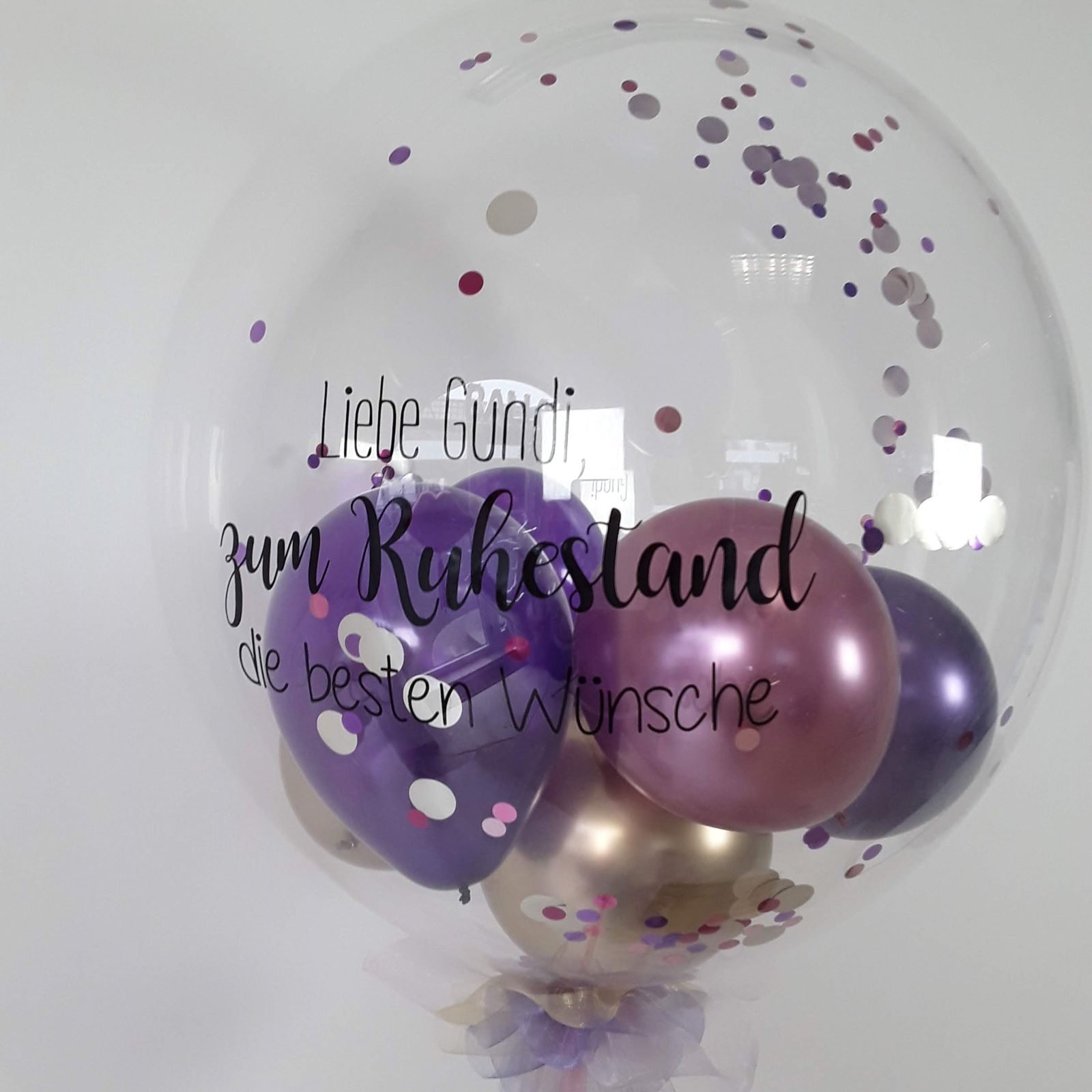 Transparenter Bubble-Ballon mit lila, fliederfarbenen und goldenen Ballons, Konfetti und schwarzem Aufdruck 'Liebe Gundi zum Ruhestand die besten Wünsche'.