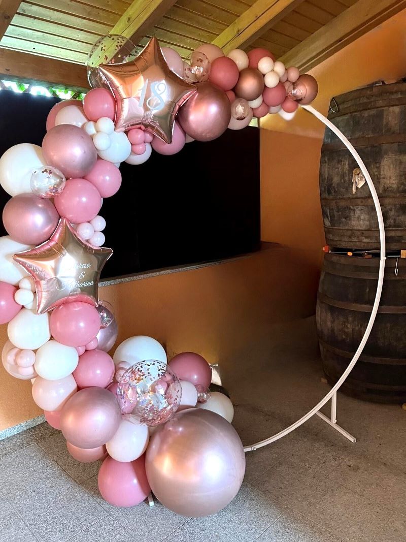 Ringbogen aus Ballons in Roségold, Rosa, Pink und Weiß, dekoriert mit großen Sternfolienballons mit Aufdruck '18' und 'Theresa&Katharina' und transparenten Konfettiballons, aufgebaut in einem Raum mit Holzdecke und zwei Holzfässen im Hintergrund