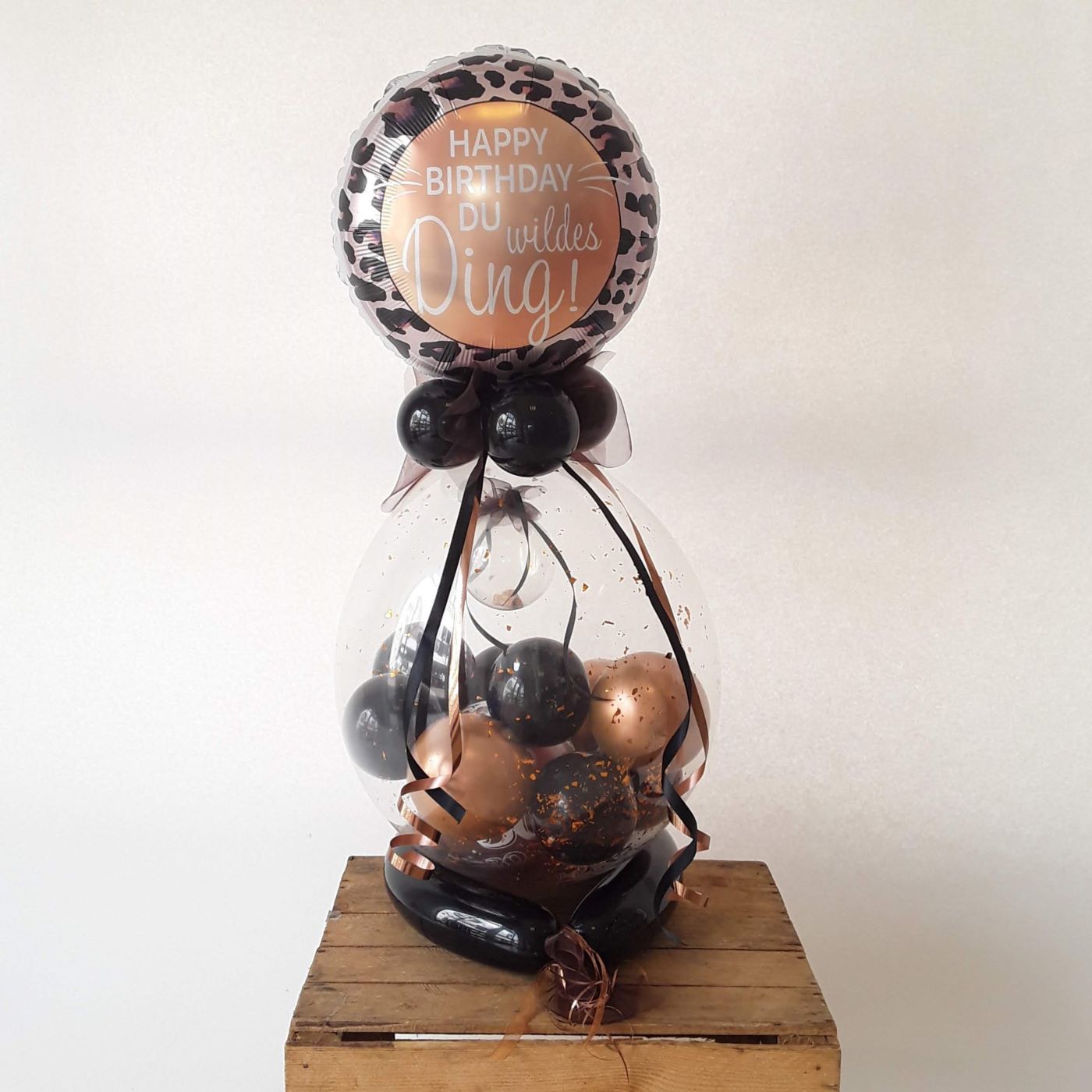 Geburtstagsballon mit Aufdruck 'Happy Birthday du wildes Ding' in Schwarz-Bronze und Leoprint, darunter ein durchsichtiger Ballon mit schwarzen und bronzenen kleinen Ballons, durchsichtiger Kugel für Geldscheine, Konfetti, verziert mit Schleifen