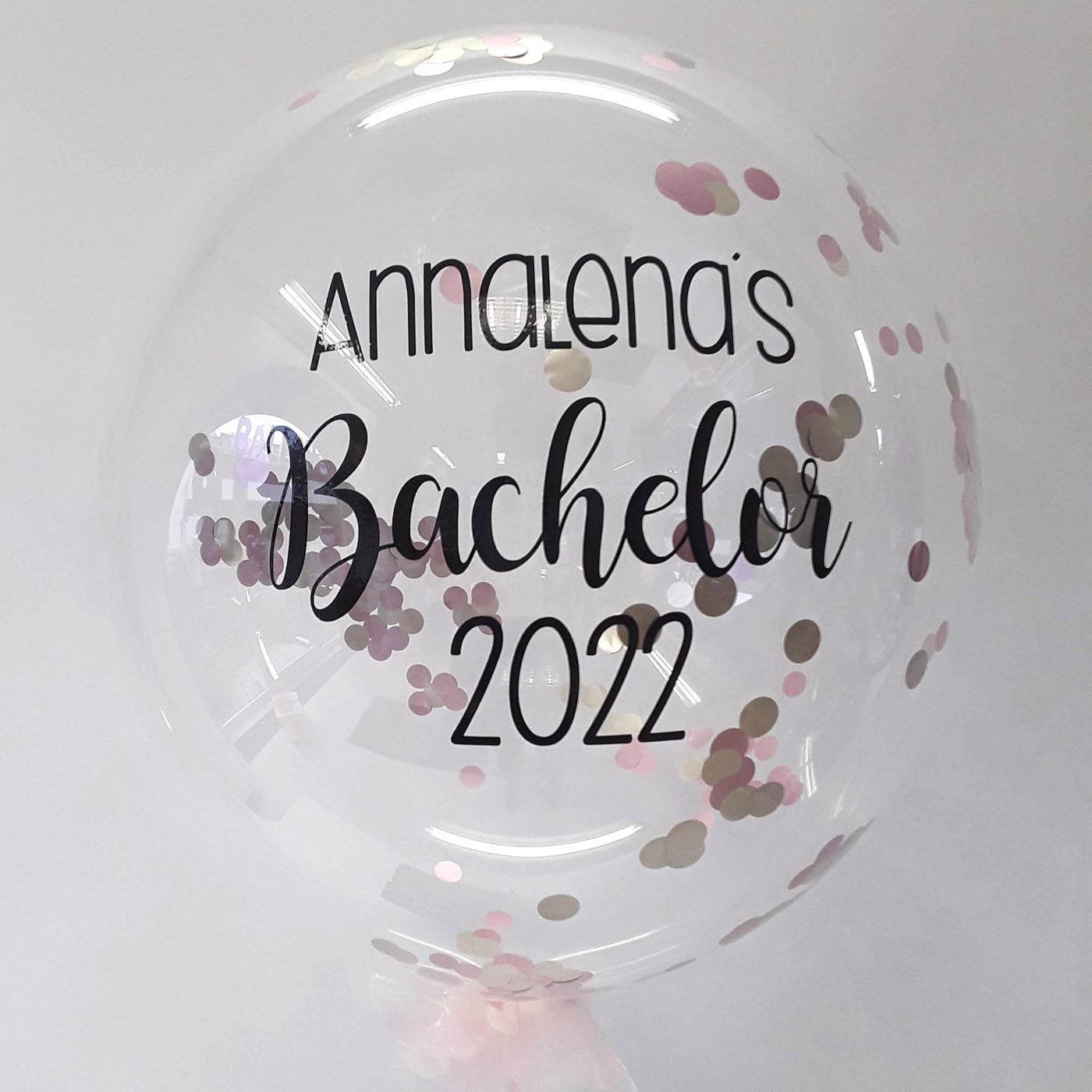 Transparenter Konfetti-Bubble mit Aufdruck „Annalena’s Bachelor 2022“. Im Inneren rosé- und goldfarbene Konfettikreise, unten zarter Tüll.