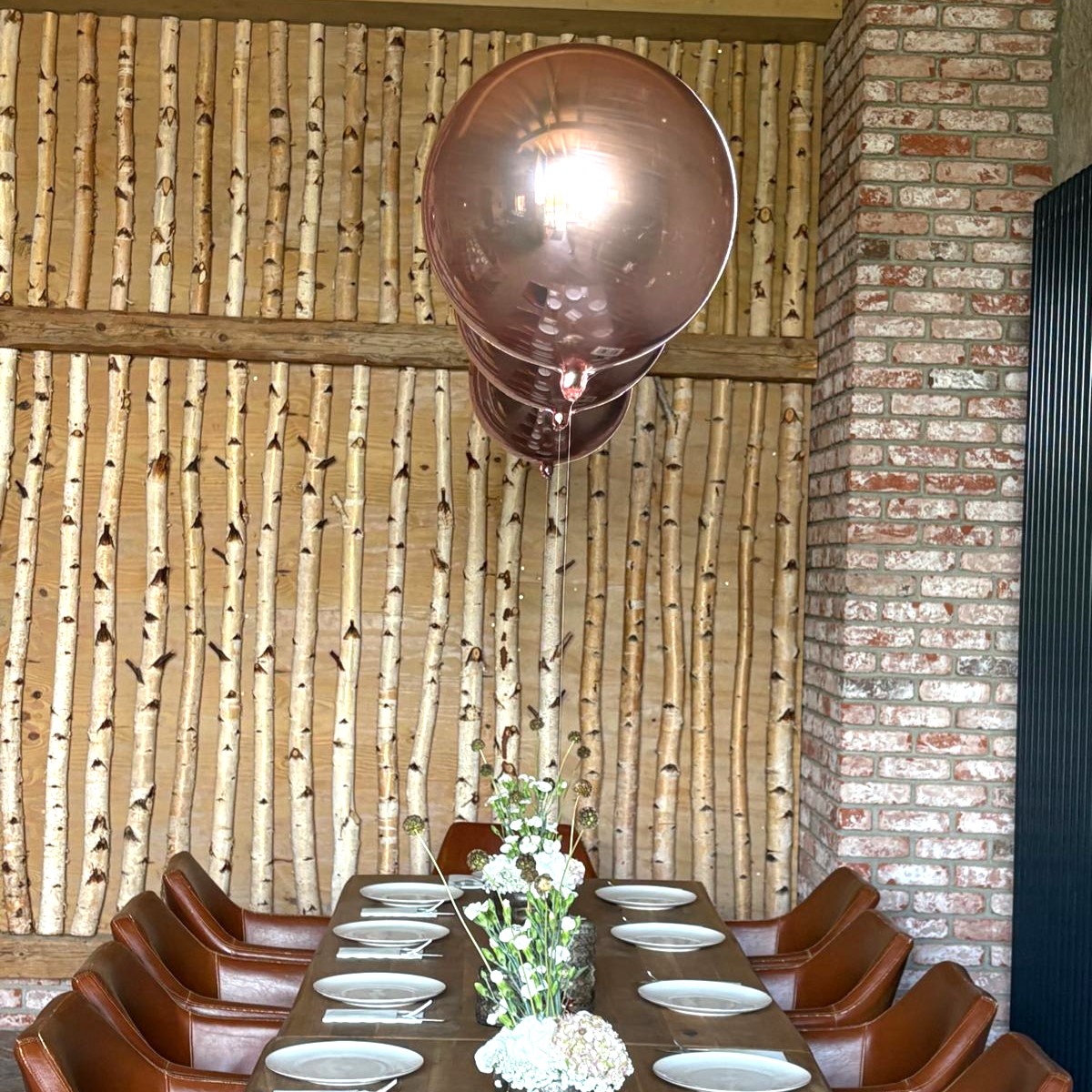 3 Schwebende Orb-Ballons in glänzendem Roségold als elegante Tischdekoration über einer festlich gedeckten Tafel in einem Raum mit Mauer- und Holzwänden