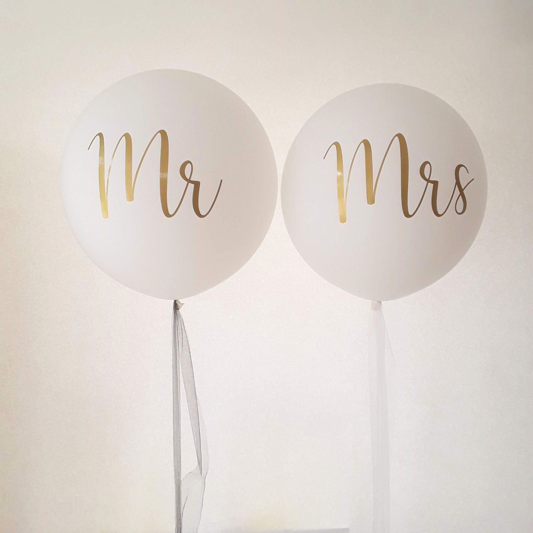 Zwei große weiße Ballons mit der goldenen Aufschrift 'Mr' und 'Mrs', jeweils mit einem langen weißen Band.