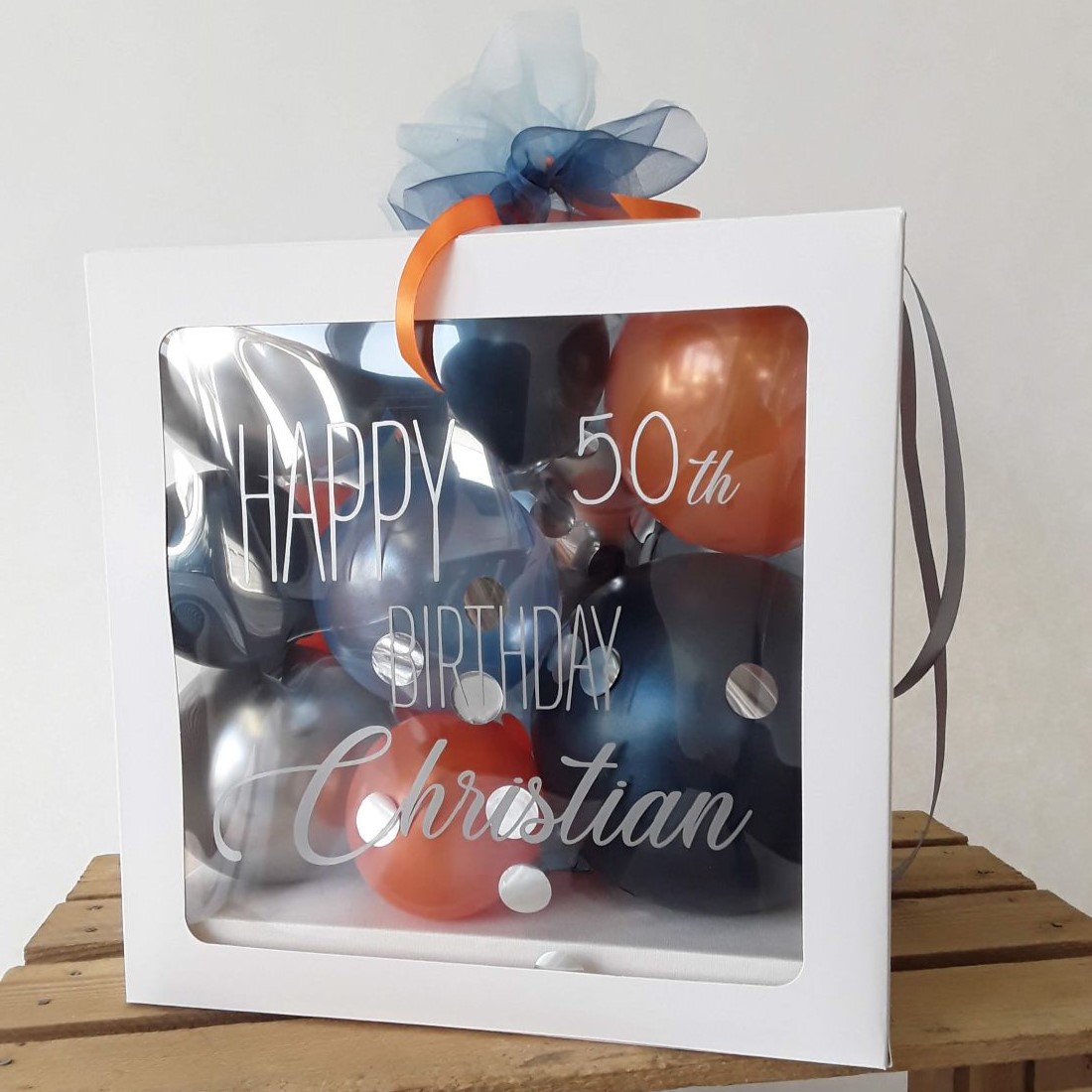 Ballonbox mit Aufdruck 'Happy 50th Birthday Christian', gefüllt mit Ballons in Blau, Silber und Orange, Schleife in Blau
