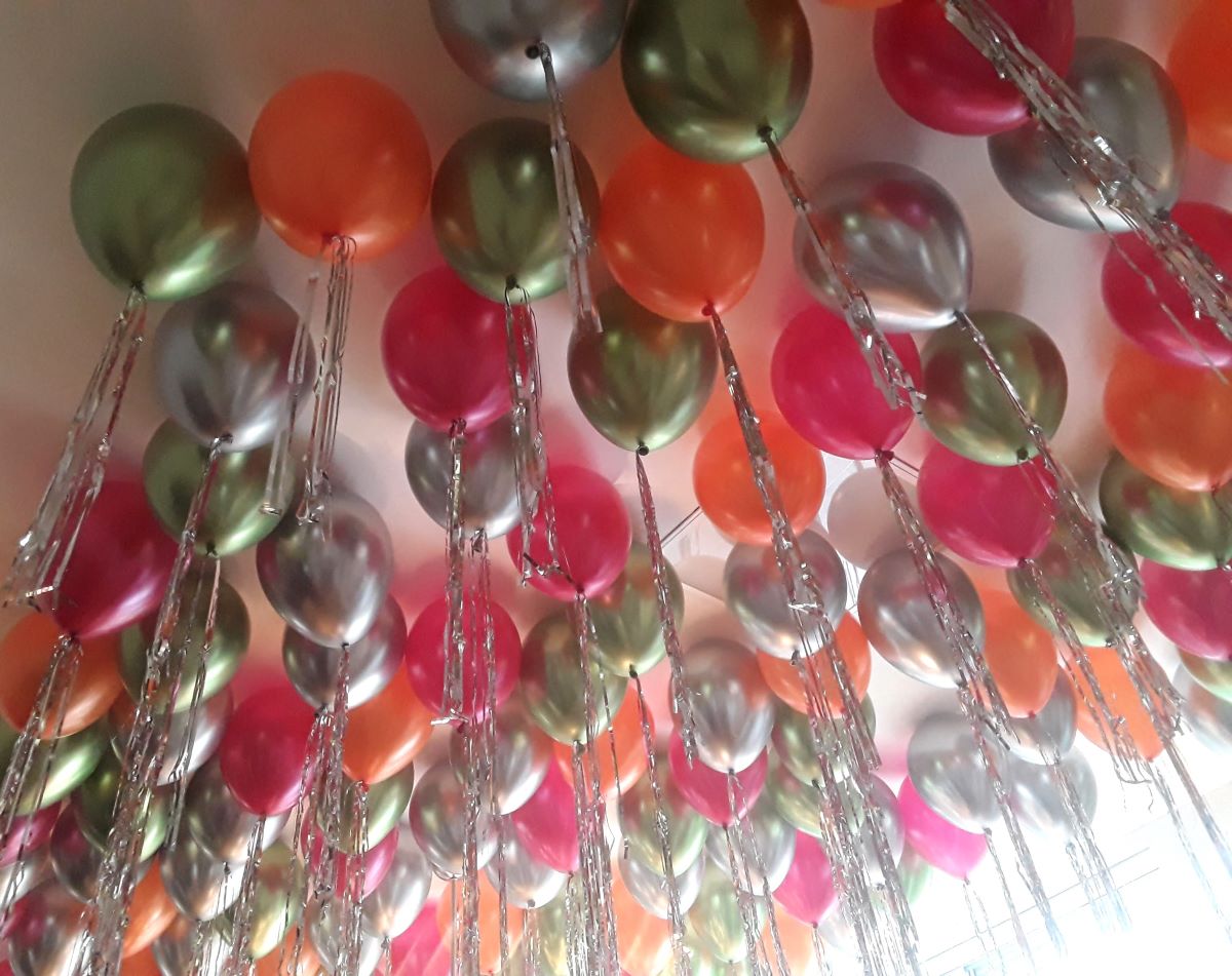 Vielzahl an Heliumballons in Pink, Orange, Grün und Silber mit metallic-glänzender Oberfläche und langen silbernen Flitterketten, schwebend an einer Zimmerdecke