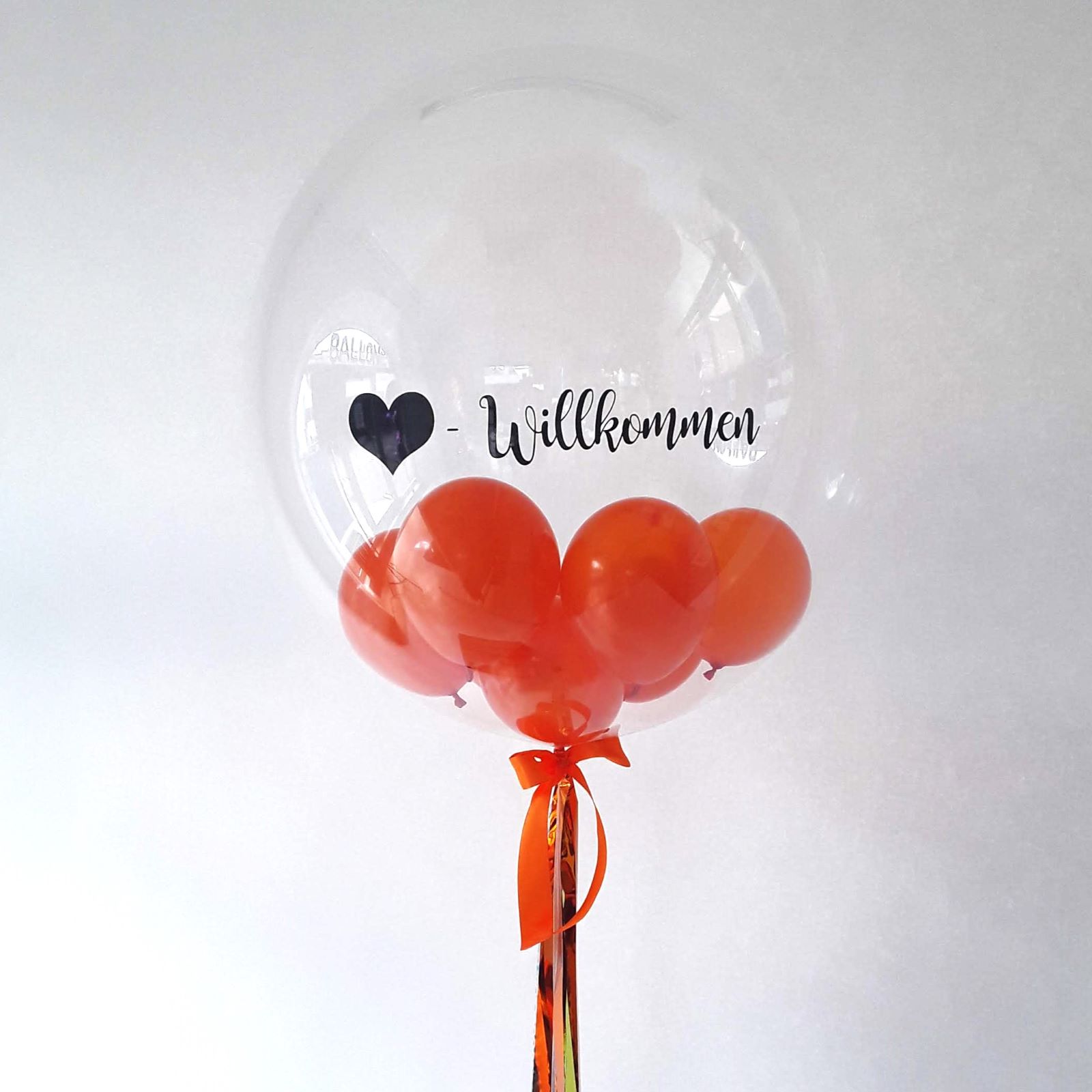 Transparenter Bubble-Ballon mit schwarzem Herz-Aufdruck und dem Schriftzug 'Willkommen', gefüllt mit mehreren orangen Mini-Ballons und dekoriert mit orangem Schleifenband.