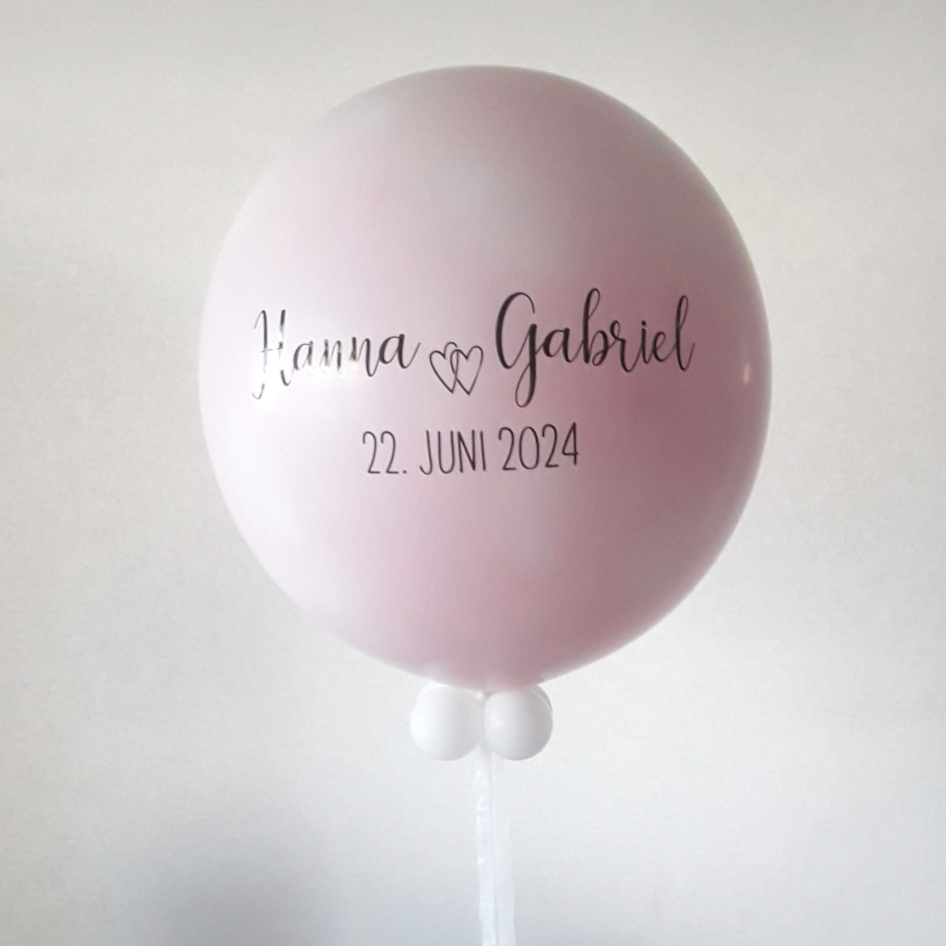Großer rosa Heliumballon mit schwarzer Aufschrift 'Hanna ♥ Gabriel 22. Juni 2024', dekoriert mit kleinen weißen Ballons am Boden.