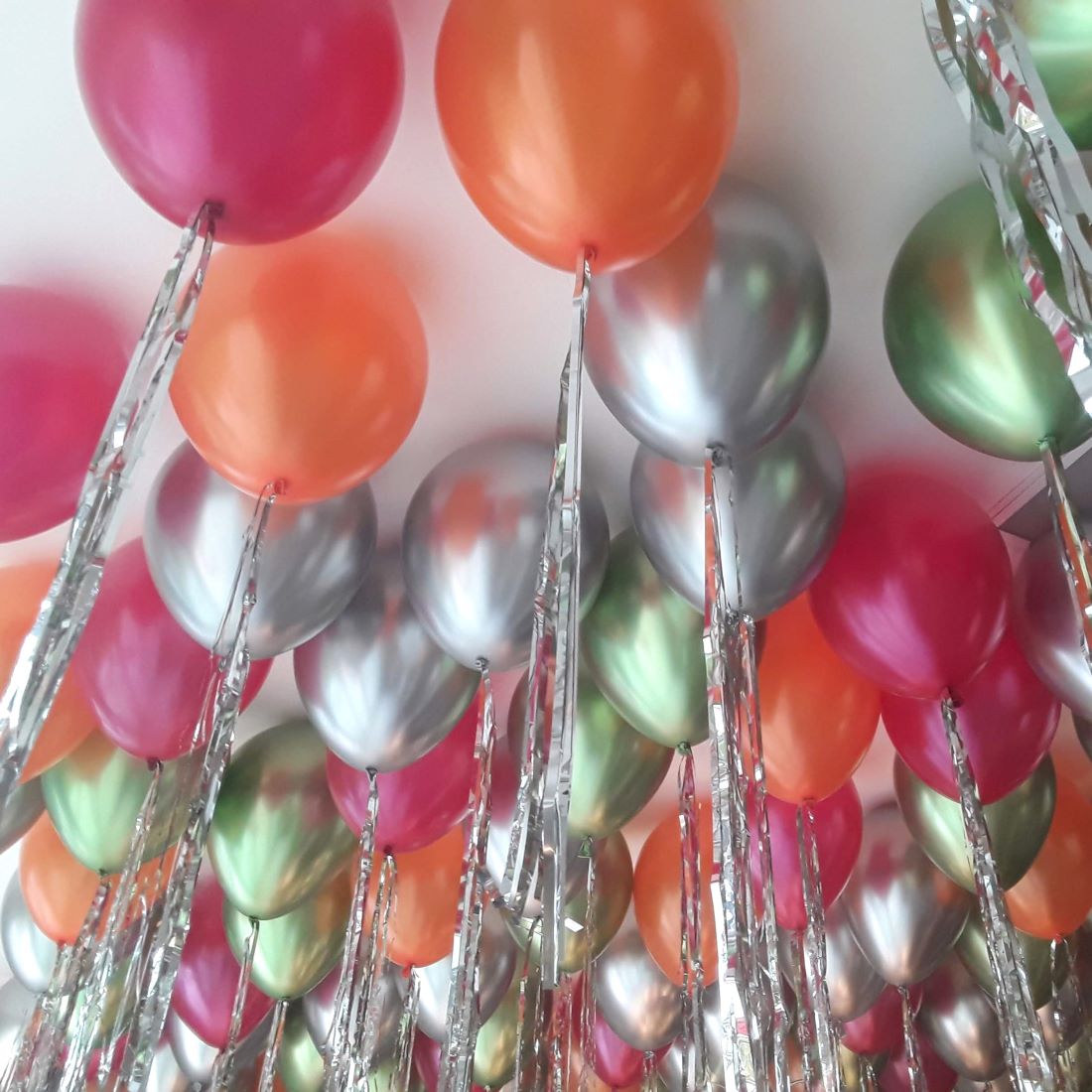 Vielzahl an Heliumballons in Pink, Orange, Grün und Silber mit metallic-glänzender Oberfläche und langen silbernen Flitterketten, schwebend an einer Zimmerdecke