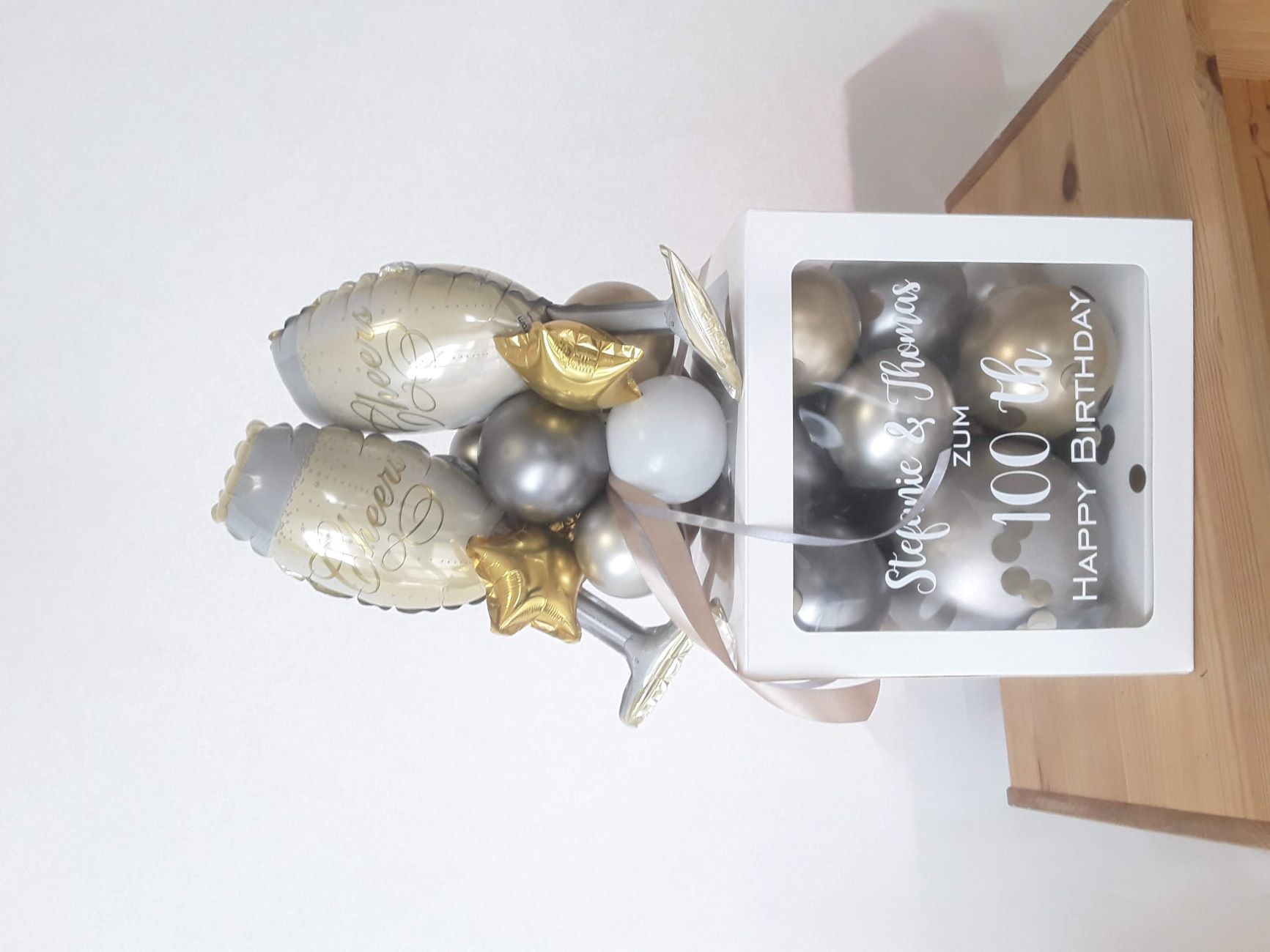 Ballonbox mit silber-goldenem Glanz, befüllt mit Ballons, zwei Sektglas-Folienballons und goldenen Sternen, Aufdruck mit Namen und Geburtstagszahl.