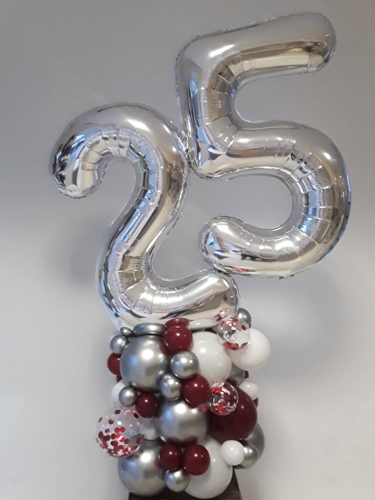 Große silberne Zahlenballons 2 und 5 auf einem dichten Sockel aus Ballons in Silber, Weiß und Bordeaux sowie transparenten Konfetti-Details.