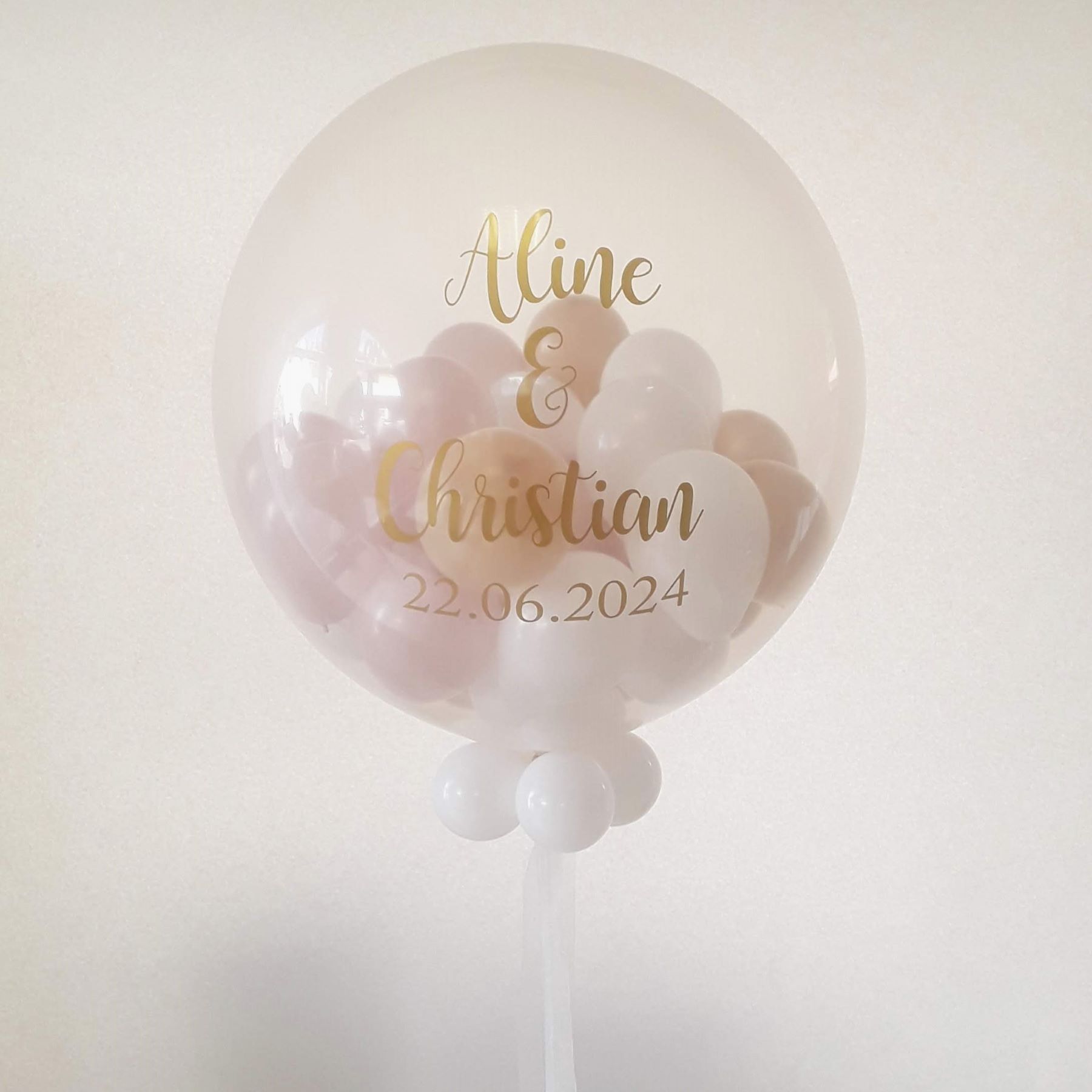Durchsichtiger Riesenballon (60 cm) gefüllt mit ca. 30 kleinen Innenballons in Pastellfarben, außen mit goldfarbigem Aufdruck „Aline & Christian, 22.06.2024“ in eleganter Schrift, dekoriert mit weißen Miniballons am Ballonhals