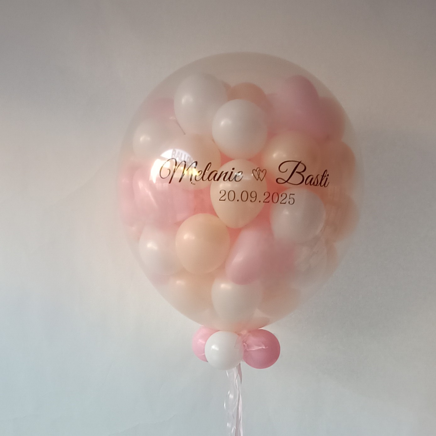 Transparenter ca.80‑cm‑Ballon mit bis zu 80 Innenballons in weiß und creme und rosa; auf dem Außenballon steht in goldener Schrift  die Namen des Brautpaars und das Datum, weißer Hintergrund